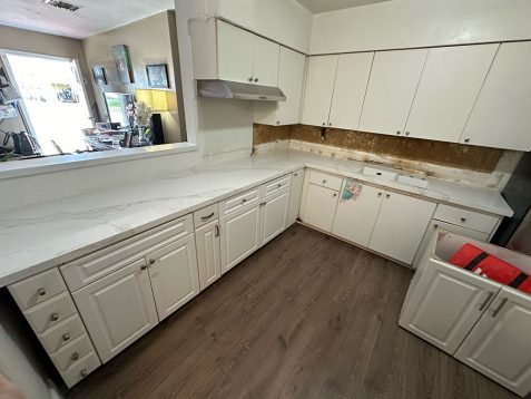 A modern kitchen with white cabinets and marble countertops, featuring an unfinished wall area and a visible red bag in the open drawer.