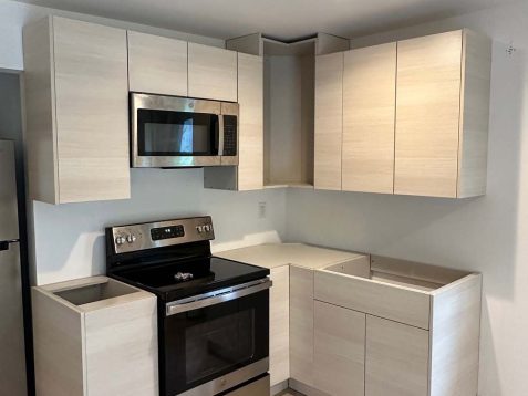Modern kitchen with light wood cabinets, stainless steel appliances, and unfinished cabinet spaces, featuring a black stove and microwave.