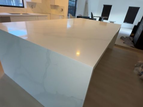 Modern kitchen featuring a large white island with subtle veining, surrounded by minimalist decor and large windows for natural light.