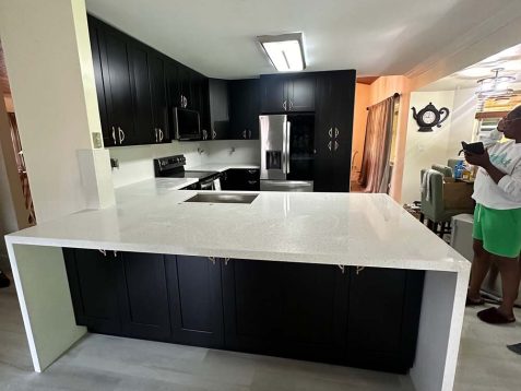 Modern kitchen with black cabinets, white quartz countertop, and stainless steel appliances, featuring a person using a mobile phone nearby.