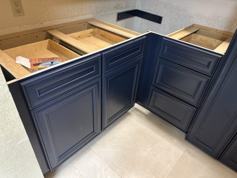 A partially assembled kitchen cabinet with blue doors and empty drawers, against a wall with unfinished drywall, on tiled flooring.