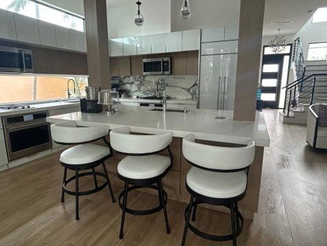 Modern kitchen featuring sleek white bar stools, marble countertops, stainless steel appliances, and an open layout with natural light.