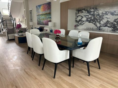 A modern dining area featuring a glass table surrounded by white chairs, with artwork and a marble kitchen backsplash in the background.
