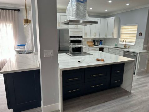 Modern kitchen featuring white cabinetry, a navy blue island, marble countertops, and stainless steel appliances, with natural light streaming in.